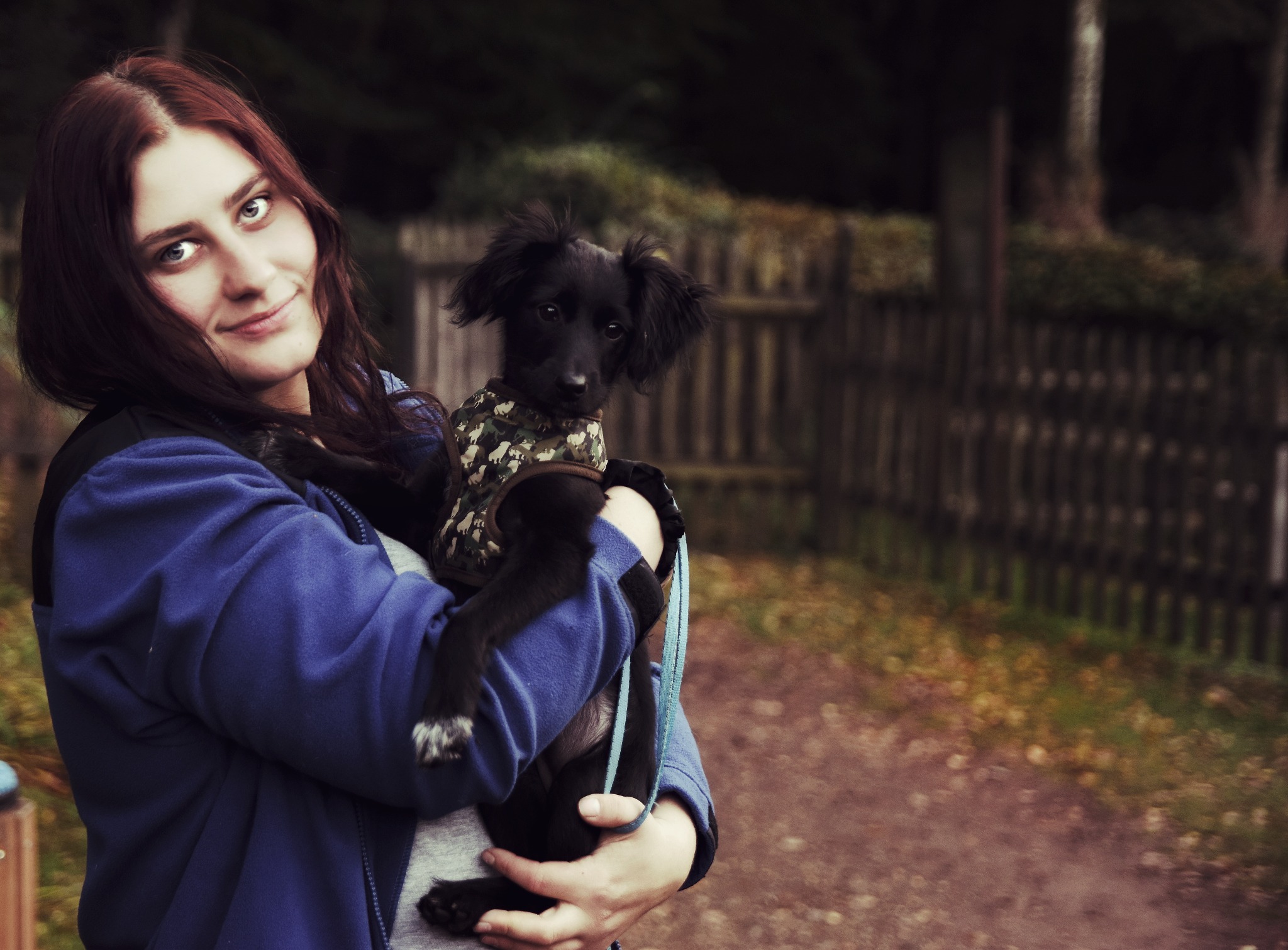 Portret kobiety z rudymi włosami trzymającej na rękach czarnego szczeniaka w ubranku w Raciborzu, na tle drewnianego płotu i ścieżki.
