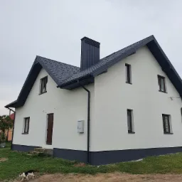 Dwukondygnacyjny dom z białą elewacją, grafitowym dachem i podmurówką, widok z boku, z kotem przechodzącym obok.