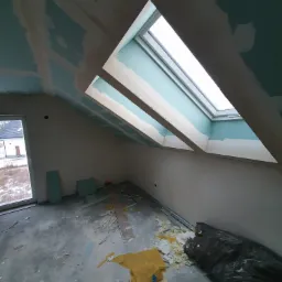 Poddasze w trakcie prac wykończeniowych: zamontowane okna dachowe, płyty gipsowo-kartonowe na skosach sufitu częściowo pokryte masą szpachlową, widoczne ślady po obróbce i resztki materiałów...