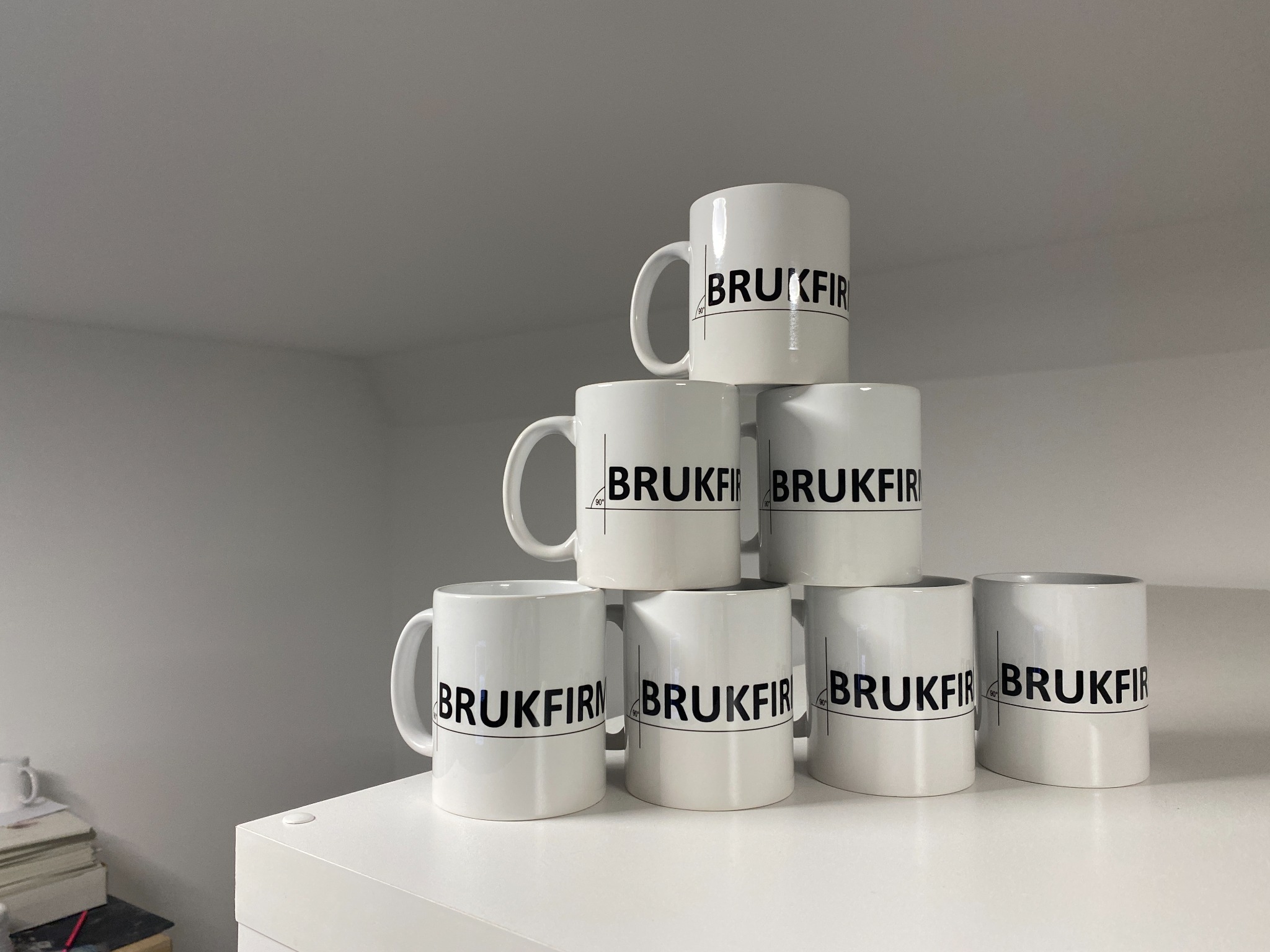 Piramida z siedmiu białych kubków z czarnym nadrukiem 'BRUKFIR' i symbolem kąta prostego, ustawiona na białej półce.