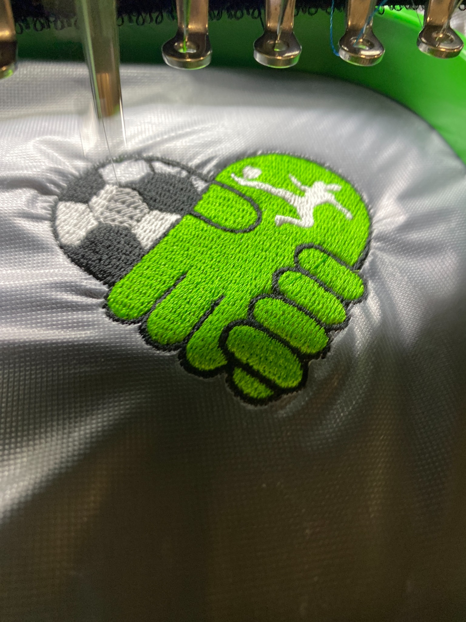 Proces tworzenia haftu z motywem piłki nożnej i zielonej dłoni z sylwetką piłkarza na szarej tkaninie, widoczne elementy maszyny hafciarskiej.