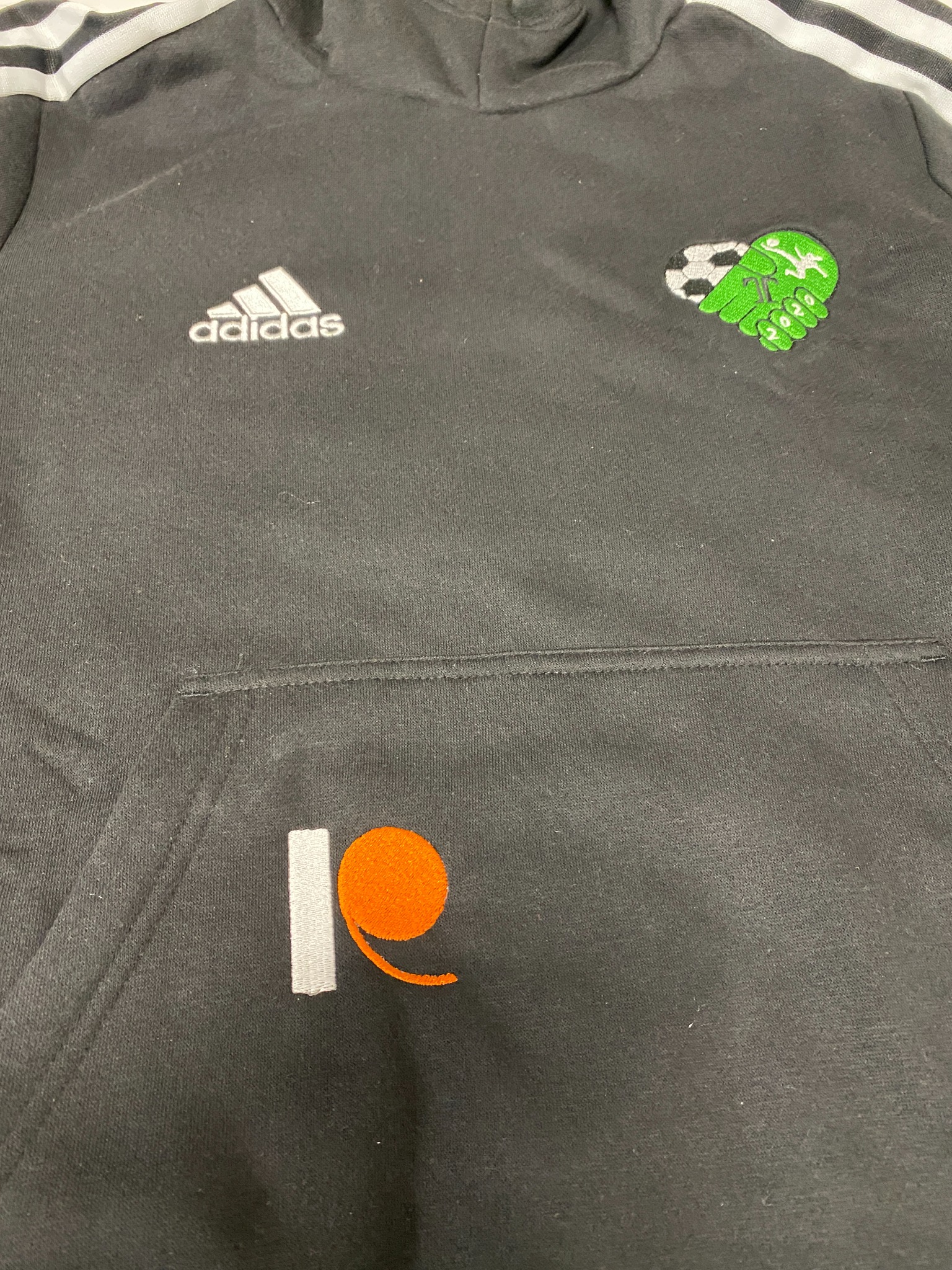 Czarna bluza Adidas z kapturem, z haftowanymi aplikacjami: logo Adidas, motyw piłkarski i pomarańczowo-biały znak graficzny na kieszeni.