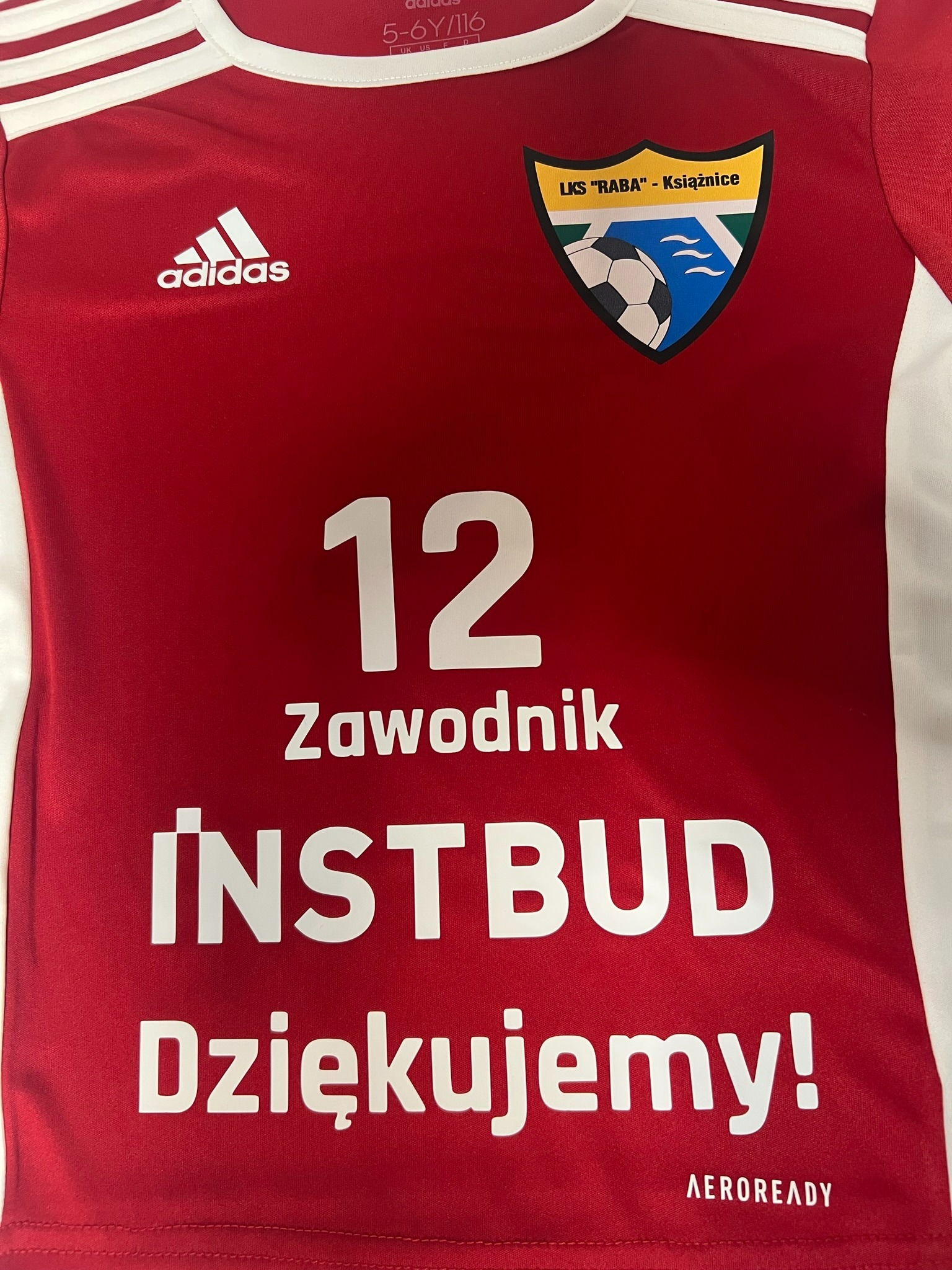 Czerwona koszulka sportowa z białymi elementami, logo Adidas, herbem LKS Raba Książnice, numerem 12, napisem 'Zawodnik', nazwą sponsora 'INSTBUD' i hasłem 'Dziękujemy!'