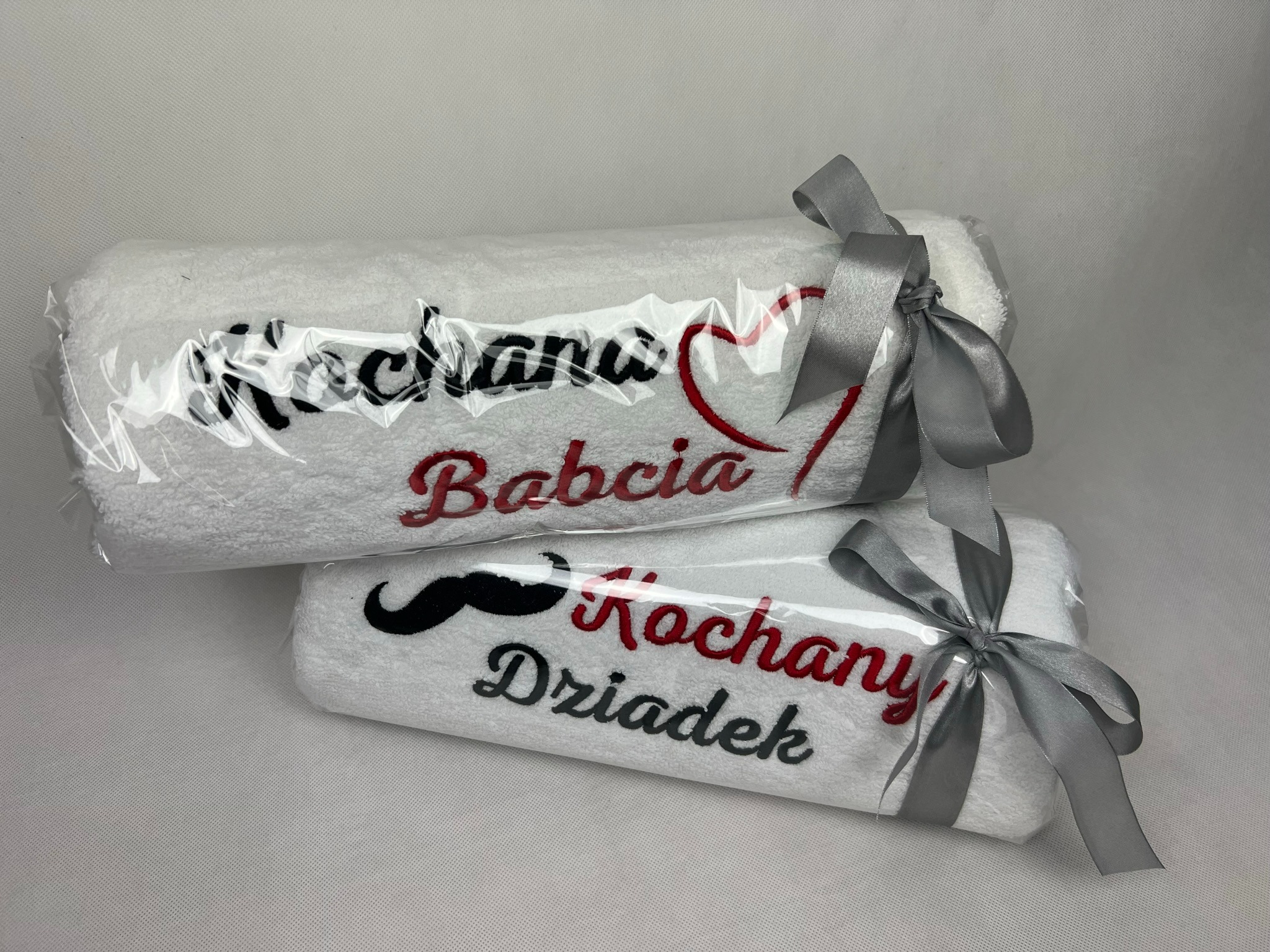 Dwa białe ręczniki frotte, każdy zapakowany w przezroczystą folię i przewiązany srebrną wstążką; na górnym ręczniku wyhaftowany napis 'Kochana Babcia' z czerwonym sercem, a na dolnym 'Kochany...