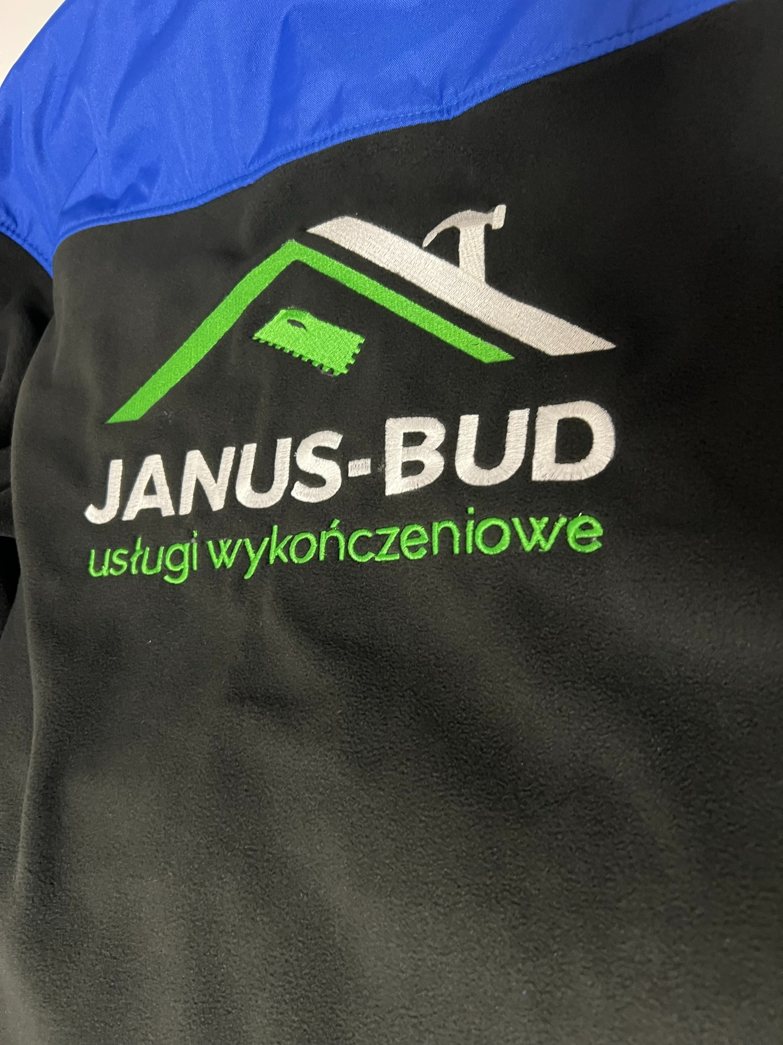 Czarna bluza polarowa z niebieskim karczkiem i wyhaftowanym logo firmy 'JANUS-BUD usługi wykończeniowe' w kolorach białym i zielonym, przedstawiającym zarys dachu, kielnię i młotek.