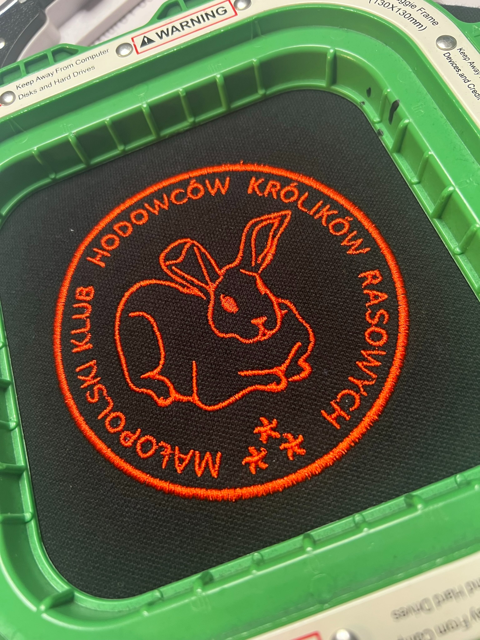 Okrągły, pomarańczowy haft z wizerunkiem królika i napisem 'Małopolski Klub Hodowców Królików Rasowych' na czarnym materiale, umieszczony w zielonej ramce hafciarskiej.