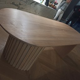 Mateusz Mrokowski Timelesswood - Owalny blat drewniany na cylindrycznej podstawie z pionowych listew, widoczny w tle fragment kuchni z marmurowym blatem i osoby.