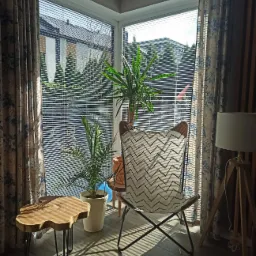 Drewniany stolik kawowy z metalowymi nogami obok fotela typu motyl z geometrycznym wzorem w pokoju z roślinami doniczkowymi i zasłonami w kwiaty.