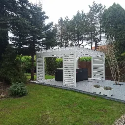 Biała pergola z żaluzjowymi ścianami na szarym tarasie, otoczona zielenią drzew i trawnikiem, z czarnym jacuzzi wewnątrz i doniczkami z ziołami na tarasie.