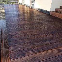 Świeżo zaimpregnowany taras z ciemnego drewna, przylegający do budynku z jasną elewacją i dwoma małymi oknami, w tle widoczne drzewa.