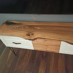 Mateusz Mrokowski Timelesswood - Szafka RTV z blatem z naturalnego drewna o nieregularnym kształcie, białe fronty szuflad z brązowymi uchwytami, metalowe nóżki, na tle drewnianej podłogi i szarej ściany.