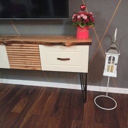 Mateusz Mrokowski Timelesswood - Stylowa szafka RTV z blatem z naturalnego drewna o nieregularnych krawędziach, białymi frontami szuflad i drewnianymi lamelami, na metalowych nóżkach typu hairpin, ustawiona na drewnianej podłodze,...