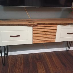 Mateusz Mrokowski Timelesswood - Biała szafka RTV z blatem z drewna o nieregularnym kształcie krawędzi, z dwoma szufladami z uchwytami ze skóry i środkową częścią z drewnianych listewek, na czarnych metalowych nóżkach.