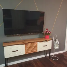 Szafka RTV z blatem z naturalnego drewna i białymi frontami szuflad, umieszczona w salonie w Nowym Dworze Mazowieckim, z widocznym telewizorem na ścianie i białym regałem w tle.