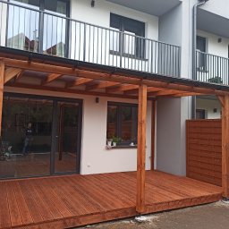 Mateusz Mrokowski Timelesswood - Nowoczesny taras z drewnianą konstrukcją zadaszenia, widok na budynek z balkonami i drewniane ogrodzenie.