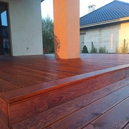 Mateusz Mrokowski Timelesswood - Ciemnobrązowy taras z desek ryflowanych, z widocznym słupem nośnym w kolorze piaskowym, fragment domu z szarą dachówką w tle, oświetlony popołudniowym słońcem.