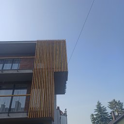 Mateusz Mrokowski Timelesswood - Nowoczesny budynek z drewnianą elewacją z pionowych listew, widziany z dołu pod kątem, na tle błękitnego nieba i okolicznych budynków.