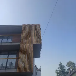 Nowoczesny budynek z drewnianą elewacją z pionowych listew, widziany z dołu pod kątem, na tle błękitnego nieba i okolicznych budynków.