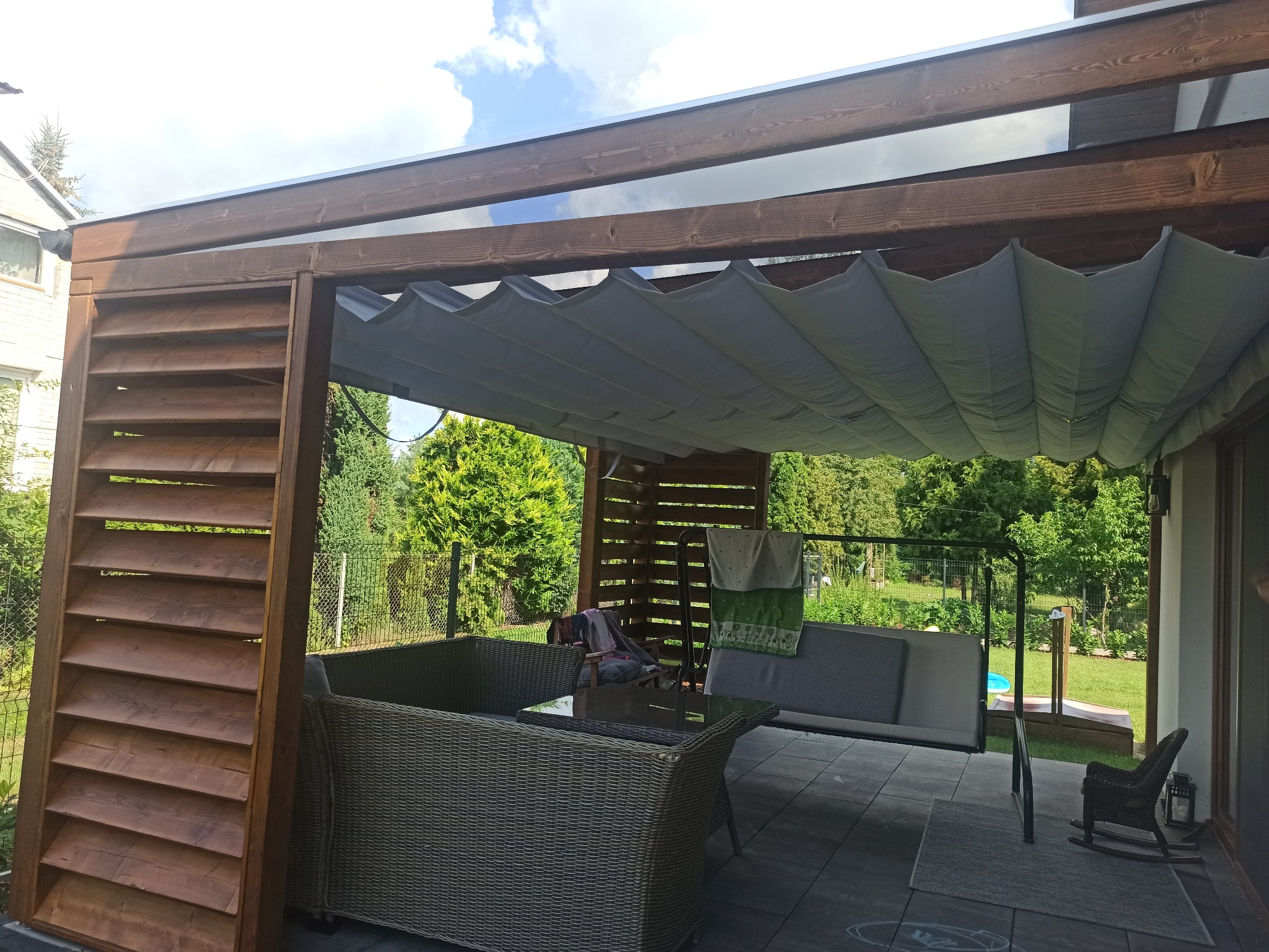 Zadaszona przestrzeń tarasowa z drewnianą konstrukcją, roletą materiałową, rattanowymi meblami ogrodowymi i huśtawką, widok na zieleń ogrodu.