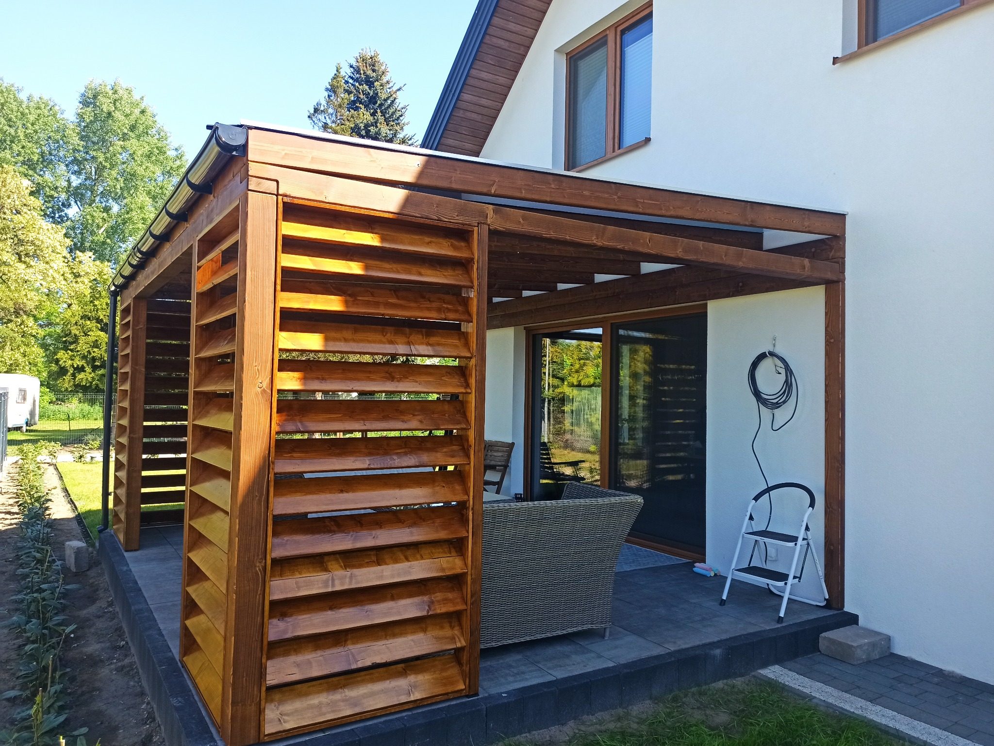 Drewniana pergola z lamelami w kolorze orzecha włoskiego, przylegająca do białego domu z widocznym tarasem i meblami ogrodowymi.