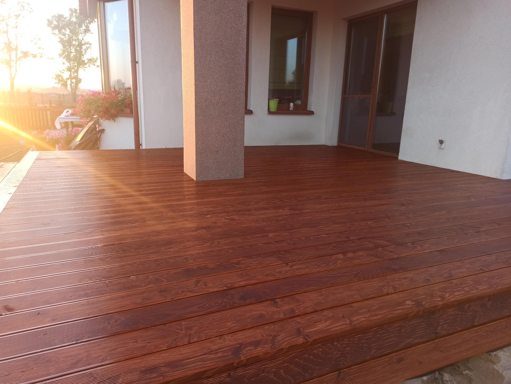 Świeżo zaimpregnowany taras z desek w odcieniach brązu, z widocznym słupem podtrzymującym zadaszenie i fragmentem domu w tle, oświetlony promieniami zachodzącego słońca.