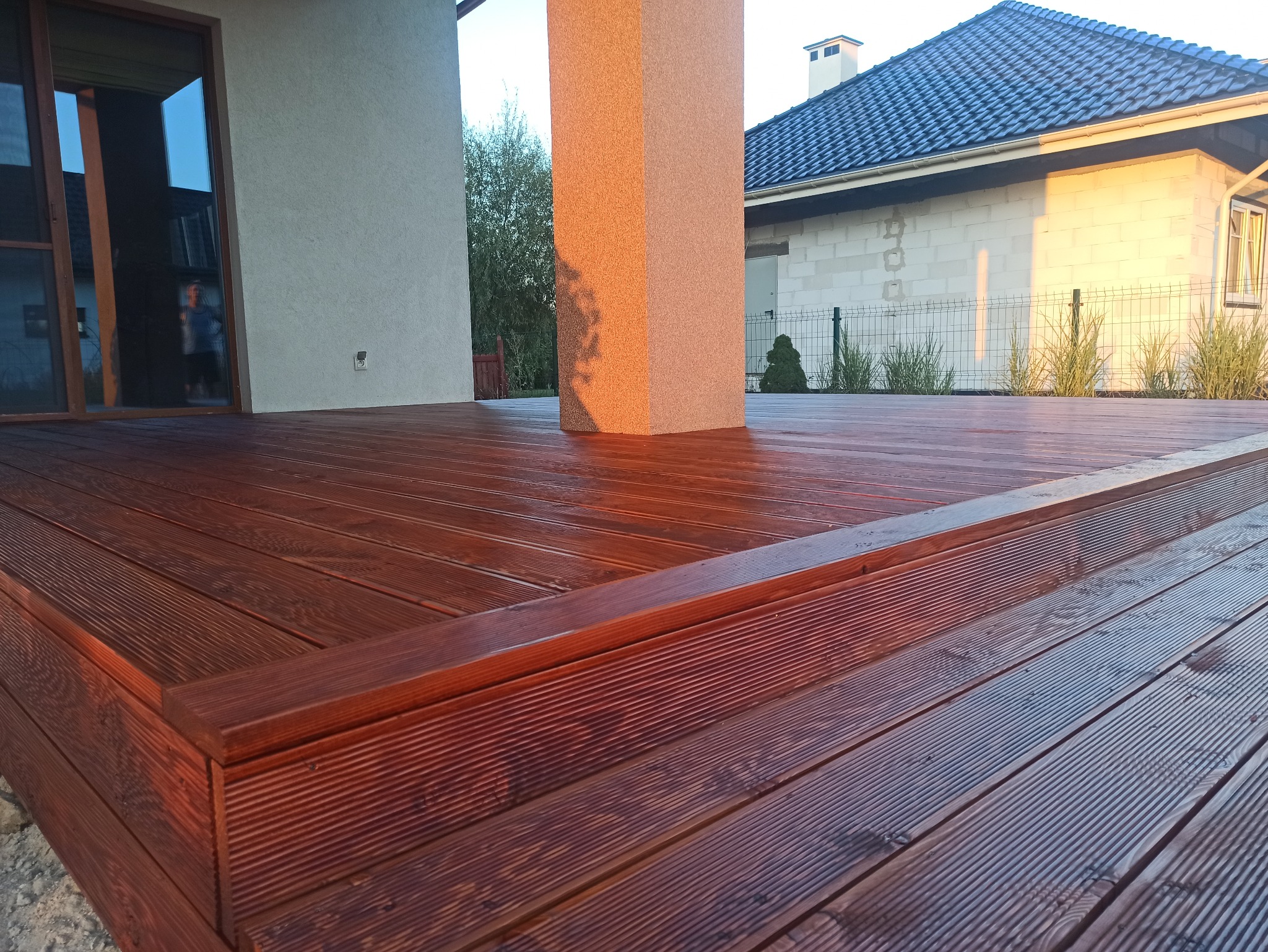 Ciemnobrązowy taras z desek ryflowanych, z widocznym słupem nośnym w kolorze piaskowym, fragment domu z szarą dachówką w tle, oświetlony popołudniowym słońcem.