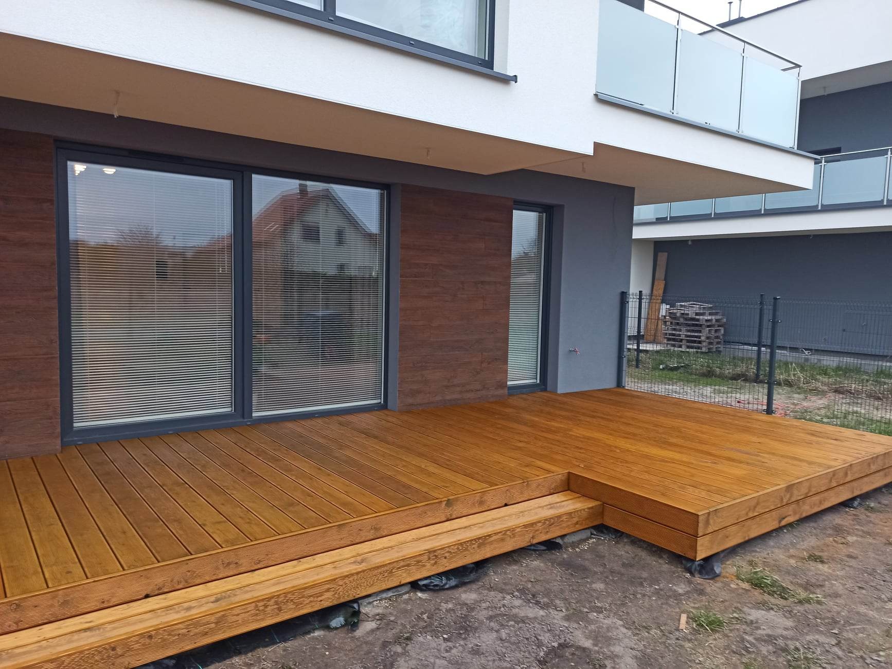 Nowoczesny taras drewniany z dwoma stopniami, widok na okno z żaluzjami i sąsiedni dom, w tle palety pod ogrodzeniem.