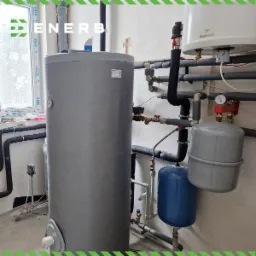 Szary, cylindryczny zbiornik na wodę w kotłowni, z widocznymi rurami, zaworami, manometrem i dwoma zbiornikami wyrównawczymi: szarym i niebieskim. W tle okno z ramą VEKA i logo Enerb w rogu.