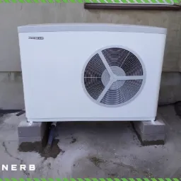 Biała jednostka zewnętrzna z wentylatorem marki Stiebel Eltron, zamontowana na betonowych blokach fundamentowych przy szarej ścianie budynku. Widoczna rura odprowadzająca skropliny. Tło stanowi...