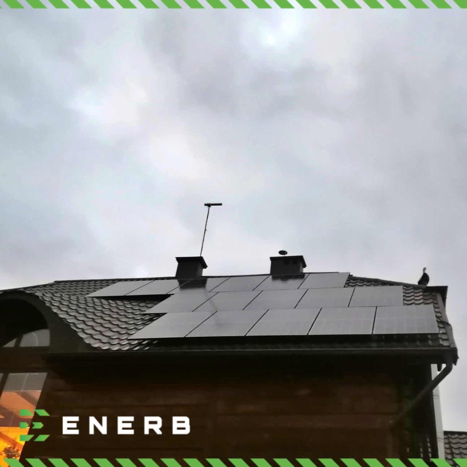 Czarny dach z brązową dachówką i panelami fotowoltaicznymi, komin, antena telewizyjna, logo ENERB na dole zdjęcia, pochmurne niebo w tle.