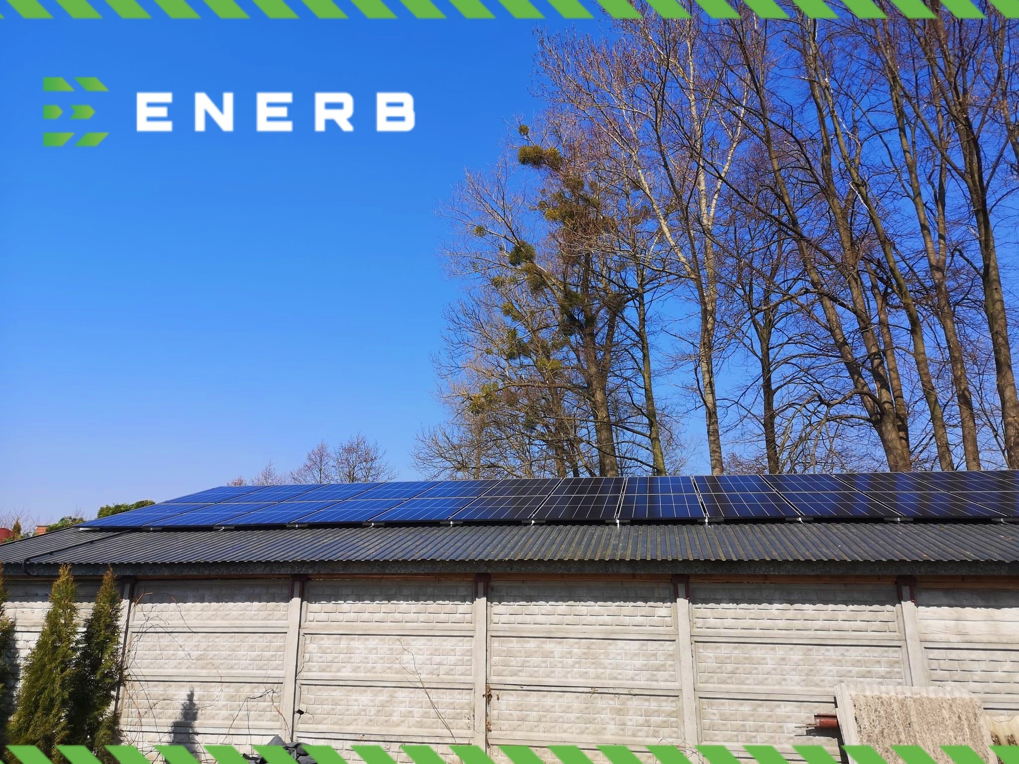 Instalacja paneli słonecznych na dachu budynku z betonowym ogrodzeniem, widoczne drzewa bez liści i błękitne niebo, logo firmy w lewym górnym rogu.