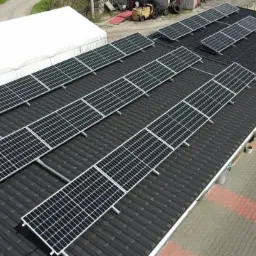 Widok z góry na instalację paneli słonecznych na dachu z czarnej blachy falistej, z widocznym fragmentem budynku z białym dachem i kostką brukową na podwórzu.