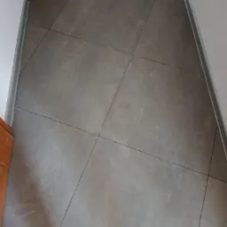 Szare, matowe płytki podłogowe ułożone w kwadraty, widoczne przy listwie przypodłogowej i krawędzi drzwi.