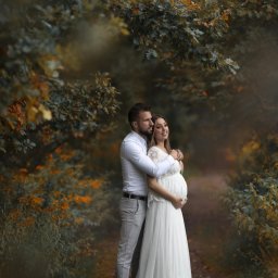 Jakub Głuszyński Usługi Fotograficzne - Przyszła mama w długiej białej sukni, obejmowana przez partnera w Tarnowie Podgórnym, na ścieżce w otoczeniu jesiennych drzew.