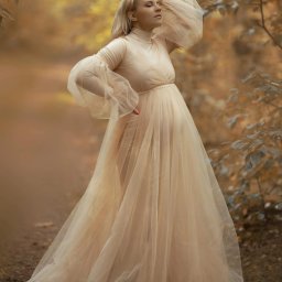Jakub Głuszyński Usługi Fotograficzne - Kobieta w ciąży w długiej, zwiewnej sukni w kolorze ecru z bufiastymi rękawami pozuje w jesiennym lesie, trzymając rękę na głowie i delikatnie się uśmiechając.