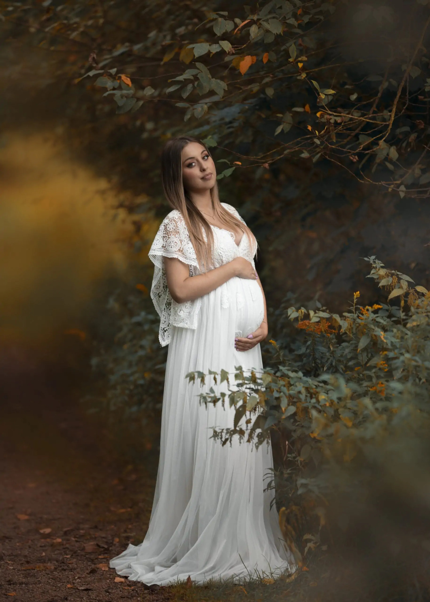 Kobieta w białej, koronkowej sukni ciążowej pozuje w plenerze, trzymając dłonie na brzuchu, z drzewami i krzewami w tle.