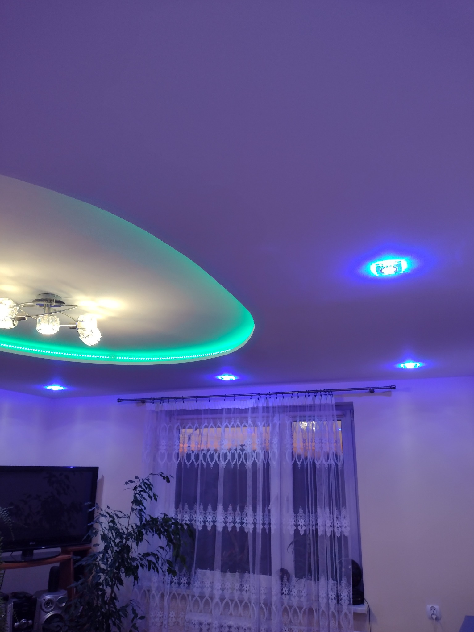 Sufit podwieszany z oświetleniem LED w kolorze zielonym i lampą wiszącą, dodatkowo punktowe oświetlenie LED w kolorze niebieskim, widok na okno z firanką i roślinę doniczkową.