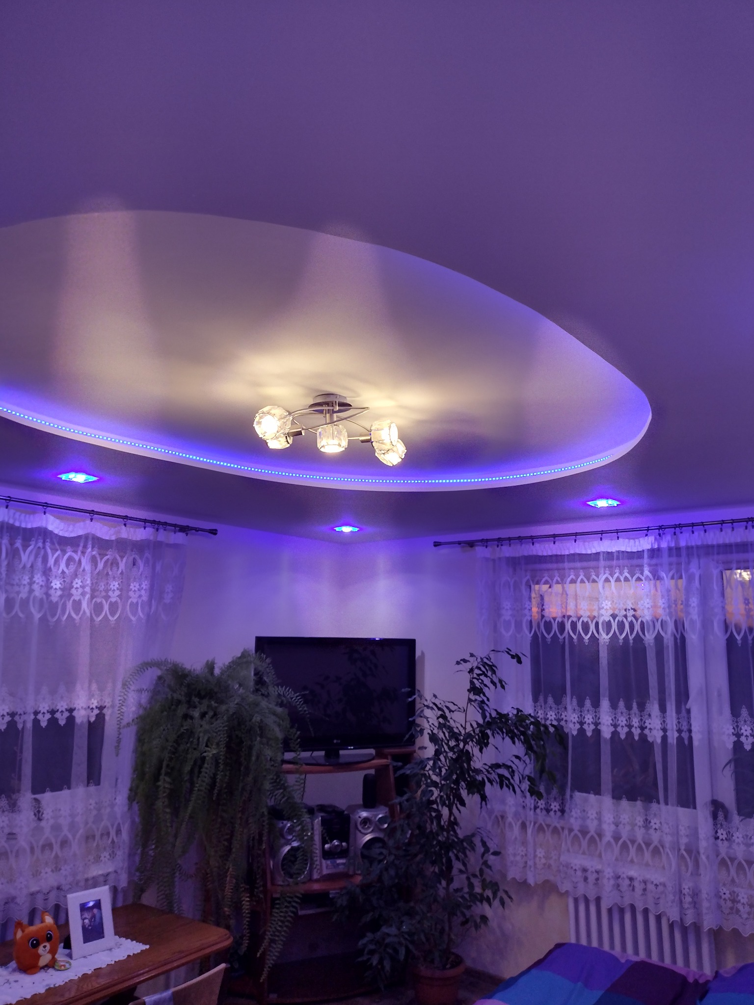 Nowoczesny sufit podwieszany z oświetleniem LED w kolorze niebieskim, widok na salon z roślinami, telewizorem i dekoracyjnymi firankami.