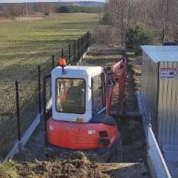Pomarańczowa mini koparka Kubota KX71-3 w trakcie wykonywania wykopu w wąskim pasie ziemi, obok metalowego garażu z tabliczką 'Uwaga! Obiekt monitorowany' i czarnego ogrodzenia panelowego.