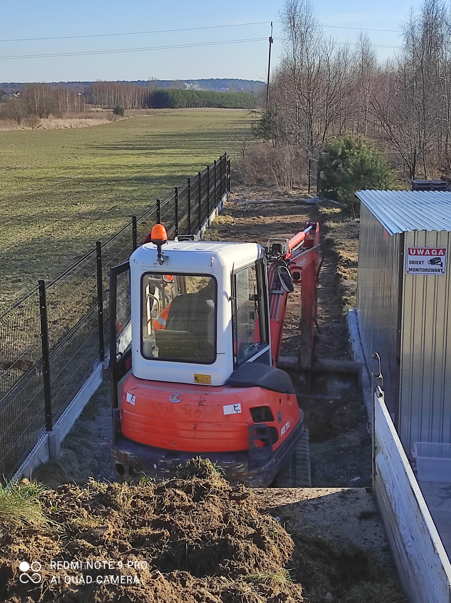 Pomarańczowa mini koparka Kubota KX71-3 w trakcie wykonywania wykopu w wąskim pasie ziemi, obok metalowego garażu z tabliczką 'Uwaga! Obiekt monitorowany' i czarnego ogrodzenia panelowego.