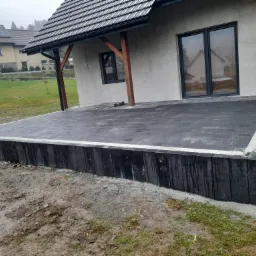 Nowo wyłożony taras z ciemnej kostki brukowej, obrzeża z jasnej kostki, widok na dom z szarym tynkiem i ciemnymi oknami, trawnik w tle.