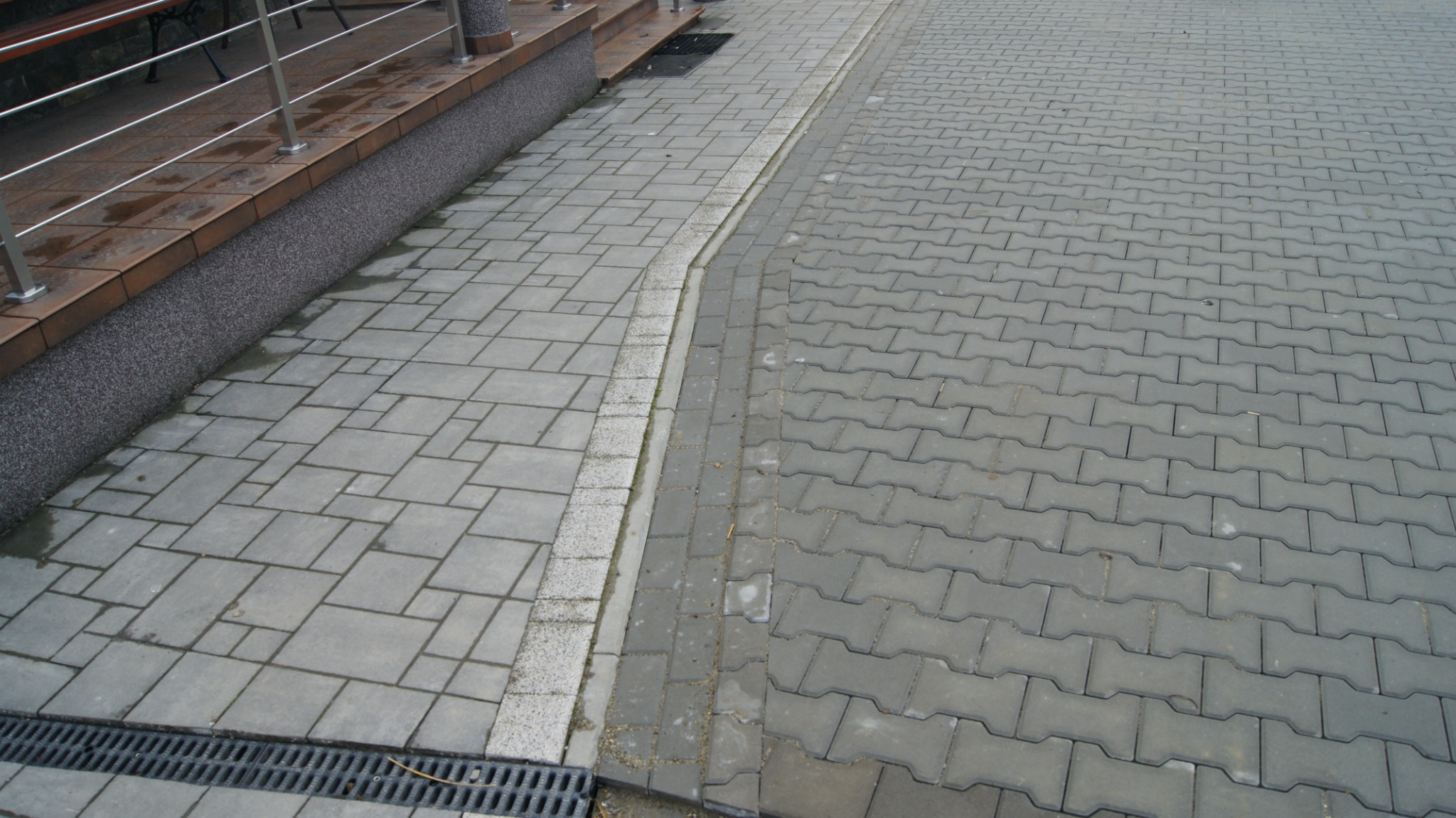Ułożona szara kostka brukowa w dwóch wzorach, oddzielona pasem z małych kostek. Widoczny fragment krawężnika i odpływ liniowy.