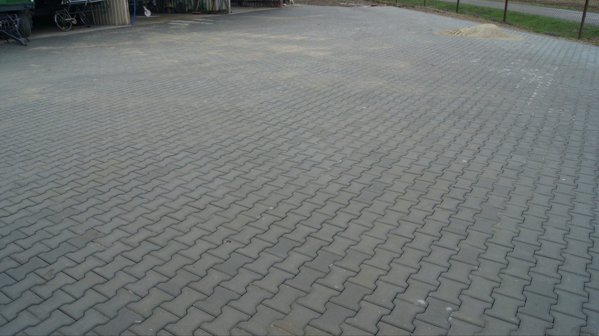 Szara kostka brukowa ułożona w jodełkę na dużej powierzchni, z widocznym stosem piasku w tle i fragmentami pojazdów. Widok z perspektywy.