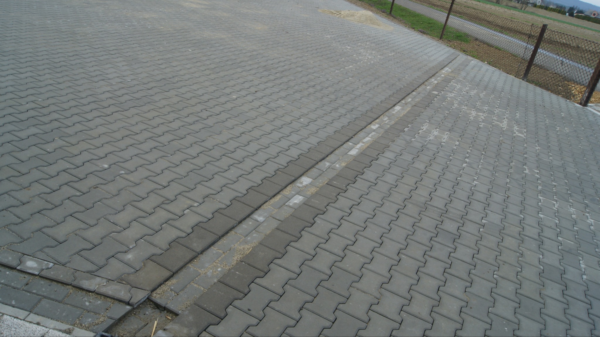 Szara kostka brukowa ułożona w jodełkę, oddzielona pasem ciemniejszej kostki. Widoczne niedociągnięcia przy krawędziach i piasek między kostkami. Tło: ogrodzenie i pole.