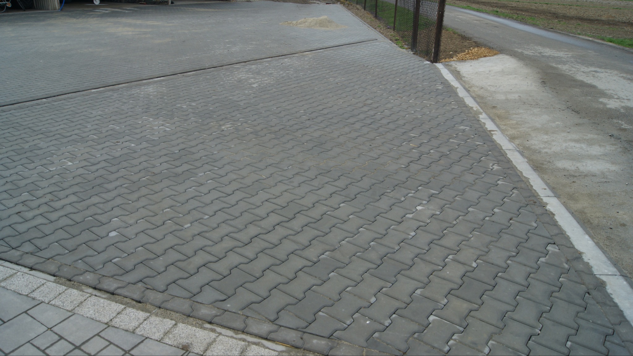 Szara kostka brukowa ułożona w jodełkę na dużej powierzchni, widoczna krawędź z jasnych kostek i fragment drogi asfaltowej z prawej strony.