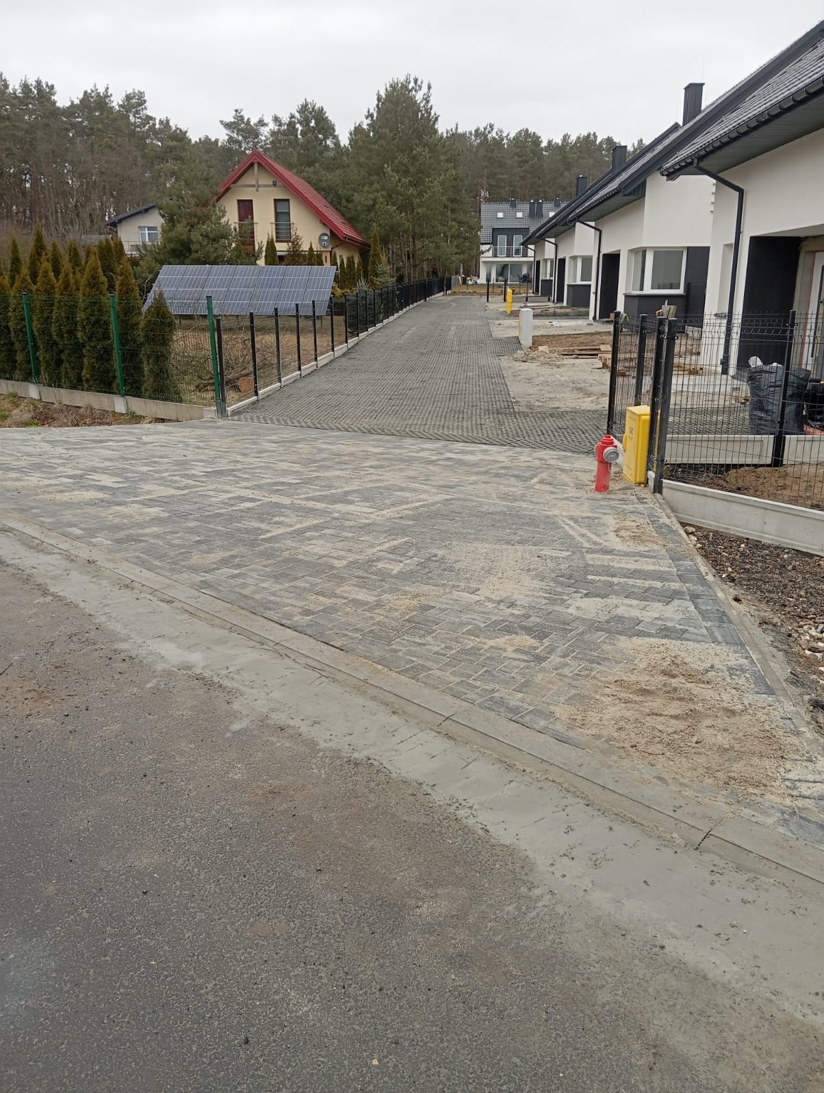 Szara kostka brukowa ułożona na podjeździe do osiedla domów jednorodzinnych, widok z poziomu ulicy, w tle drzewa i domy z panelami słonecznymi.