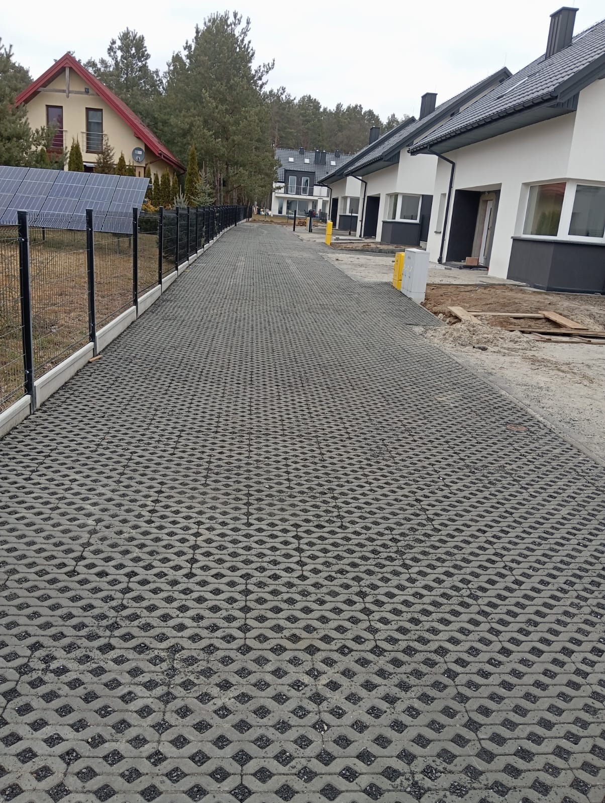 Długa, szara droga z ażurowych płyt betonowych prowadzi wzdłuż ogrodzenia i szeregu domów. Widoczne panele słoneczne i elementy wykończeniowe budynków.