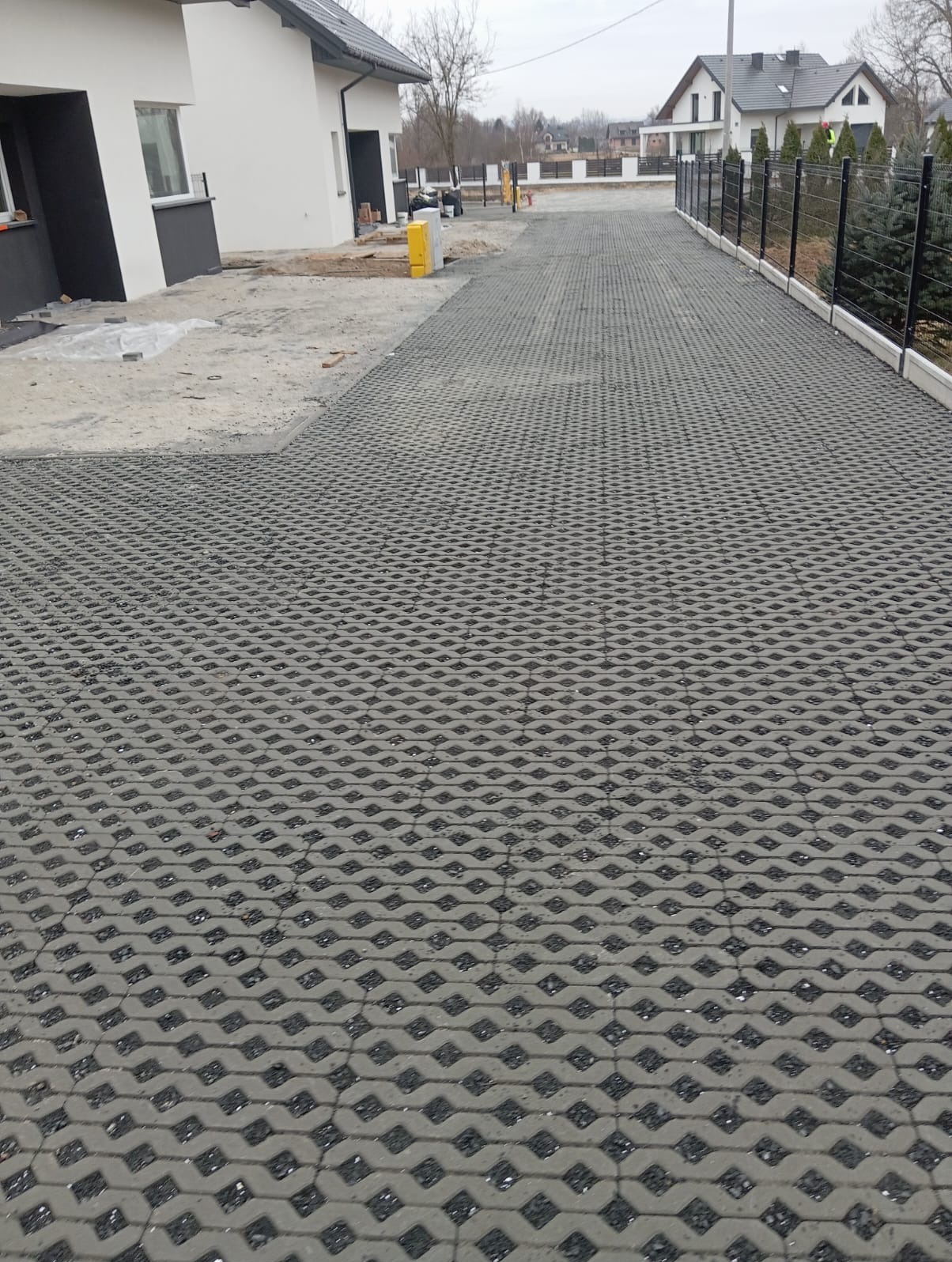 Szara, ażurowa kostka brukowa na podjeździe do domu jednorodzinnego. Częściowo ułożona, widoczne przygotowania do dalszych prac. Nowoczesne ogrodzenie w tle.