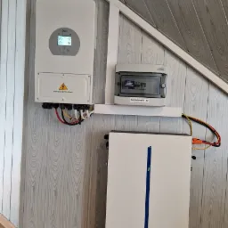 Wnętrze pomieszczenia z zamontowanym inwerterem, akumulatorem i rozdzielnicą PV-AC na ścianie wykończonej panelami imitującymi drewno.