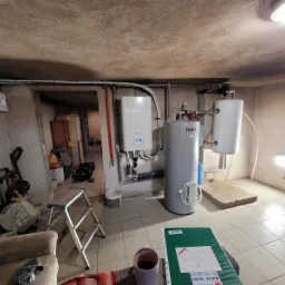 Piwnica z zamontowanym piecem gazowym, bojlerem i instalacją hydrauliczną, widoczny również fotel, drabinka i bałagan.